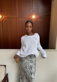 Vit kabelstickad tröja med ribbade ärmar, i kombination med en silverfärgad paljettkjol. Håller en genomskinlig kopp med iced coffee. Träbakgrund.