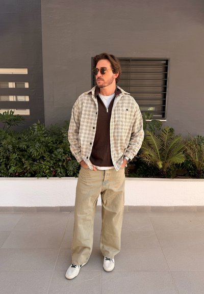 Hombre con gafas de sol, chaqueta de cuadros beige sobre suéter de cremallera marrón oscuro, pantalones beige y zapatillas blancas, de pie con las manos en los bolsillos al aire libre.