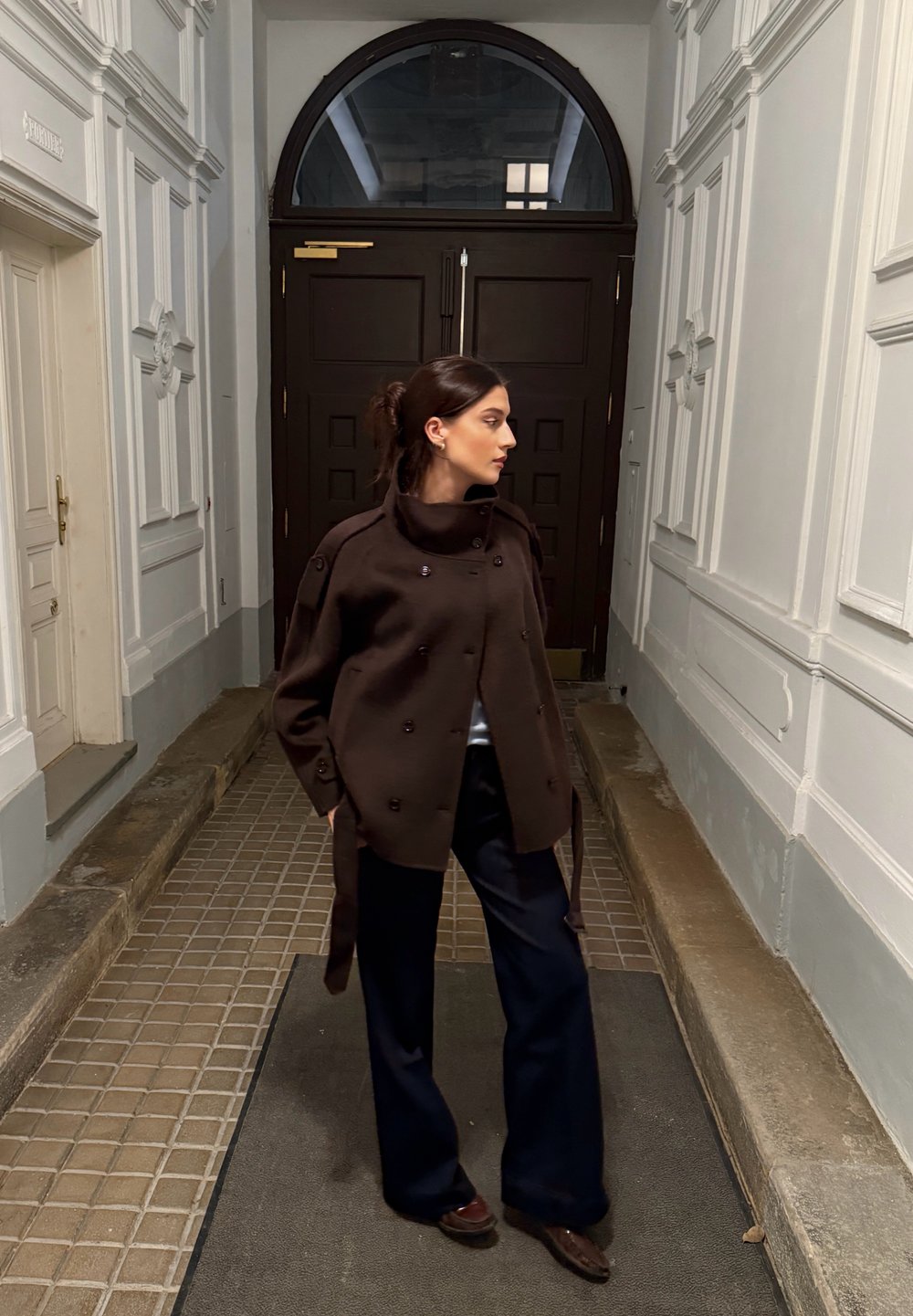 Brązowy dwurzędowy płaszcz z wysokim kołnierzem, noszony na jasnej bluzce, zestawiony z granatowymi szerokimi spodniami i ciemnymi butami, w korytarzu.