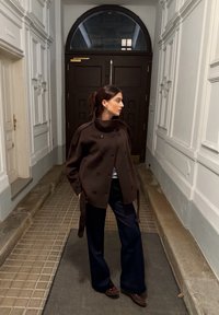 Brązowy dwurzędowy płaszcz z wysokim kołnierzem, noszony na jasnej bluzce, zestawiony z granatowymi szerokimi spodniami i ciemnymi butami, w korytarzu.