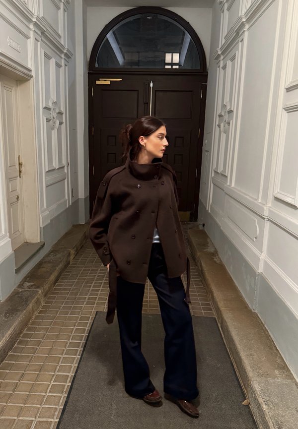 Brun dobbeltradet frakke med høj krave, båret over en lys top, parret med marineblå bukser med brede ben og mørke sko, i en korridor.