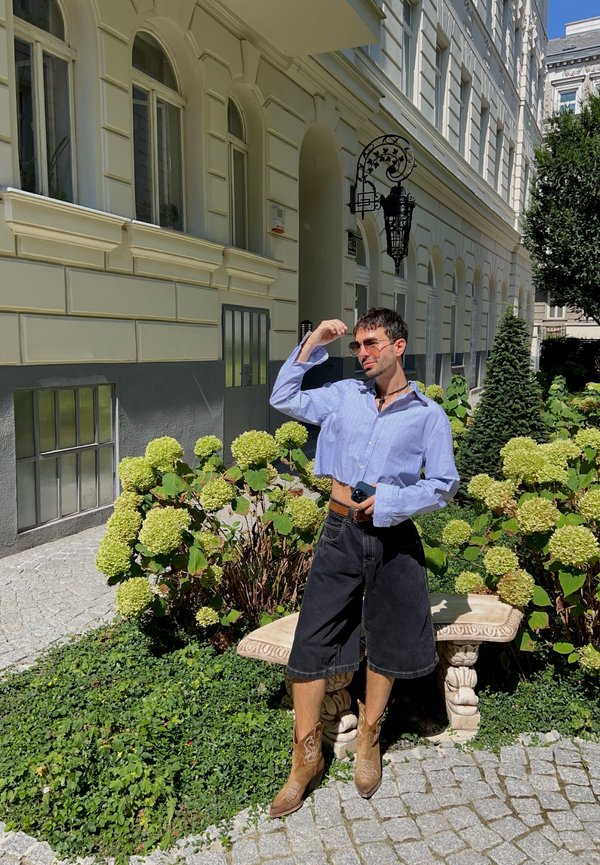 Camicia corta a strisce blu, pantaloncini di jeans neri, stivali da cowboy marroni con cuciture, in posa in un giardino vicino a ortensie verdi e panchine di pietra.