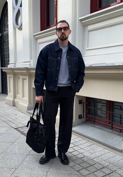 Hombre con chaqueta de mezclilla negra, sudadera gris, pantalones y zapatos negros, sosteniendo un bolso de mano negro, de pie en una acera de la ciudad junto a un edificio.