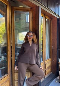 Ruskea vetoketjullinen neulepusero pitkillä hihoilla, yhdistetty ruskeisiin ribbineulottuihin leveälahkeisiin housuihin. Taustalla puiset ovet ja luonnonvalo.