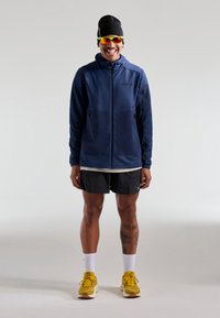 Marinsblå zip-up jacka med två framfickor, tillsammans med svarta shorts och gula löparskor. Modellen har på sig solglasögon och en svart beanie.