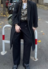 Person trägt einen dunklen Mantel, kariertes Hemd, weite schwarze Hose und schwarze Loafers und sitzt auf weißen und roten Absperrungen in einer von Bäumen gesäumten städtischen Straße.