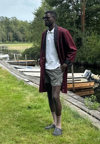 Túnica larga color burdeos sobre una camiseta polo blanca, pantalones cortos gris claro, calcetas marrones y crocs negros. De pie sobre el césped cerca de un cuerpo de agua y botes.