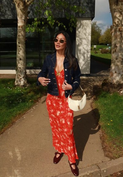 Mujer con vestido rojo de flores, chaqueta vaquera, gafas de sol y zapatos burdeos caminando por un sendero entre árboles con un bolso blanco.