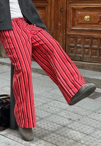 Κόκκινες και λευκές ριγέ παντελόνες με φαρδιά πόδια και κορδόνι στη μέση, συνδυασμένες με γκρι υποδήματα, τοποθετημένες σε φόντο με υφή από κυβόλιθο.