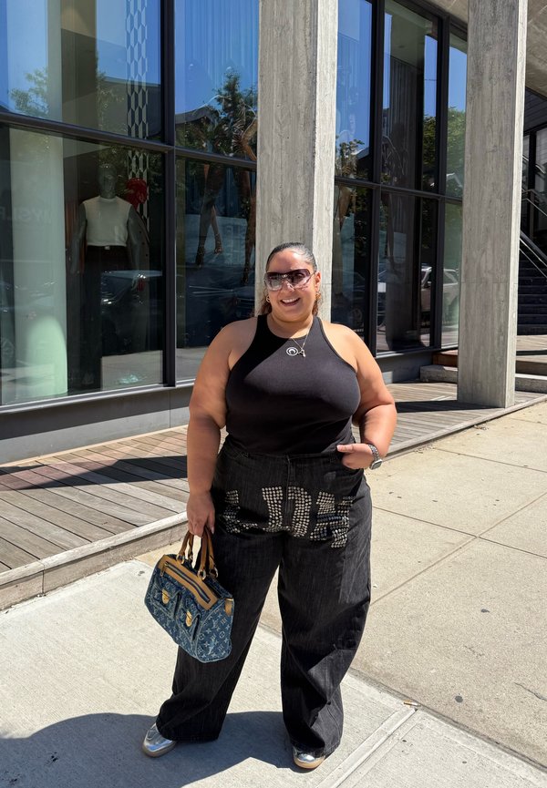 Frau trägt schwarzen ärmellosen Oberteil und weite schwarze Hose, hält eine Handtasche und steht auf dem Bürgersteig einer Stadt vor einem Glasgebäude.