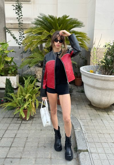 Chaqueta de cuero roja y negra con un diseño ajustado, cremalleras plateadas y una camiseta negra. Botas de plataforma negras y un bolso blanco completan el conjunto.