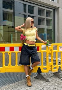 Keltainen olkapään yli menevä lyhythihainen toppi, mustat farkkibermuda-shortsit, keltainen vyö, ruskeat saappaat, pitämässä kukkakassia, jossa on vaaleanpunaisia ruusuja, aurinkolasit.