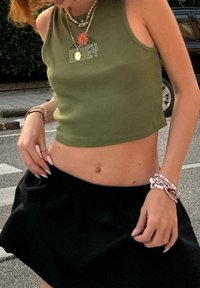 Ελαιόχρωμο crop top με την επιγραφή "OBEY", σε συνδυασμό με μια μαύρη φούστα με πτυχώσεις. Τα κοσμήματα περιλαμβάνουν βραχιόλια και ένα στρωτό κολιέ.