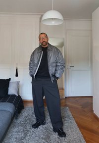 Prillidega mees, kes kannab halli bomber-jope, musta kampsunit ja halli laia pükstega, seisab vaibal moodsas elutoas.