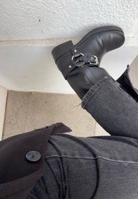 Black leather ankle boots with a rounded toe, silver buckle detail, and a low block heel, worn with dark grey jeans.