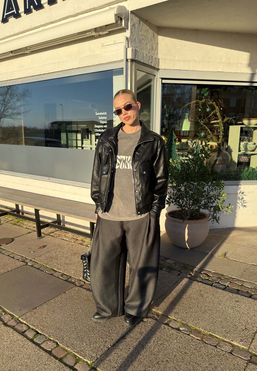 Veste en cuir noire, t-shirt graphique gris, pantalon ample foncé et chaussures noires. Lunettes de soleil fines et sac à main texturé. Cadre urbain décontracté.