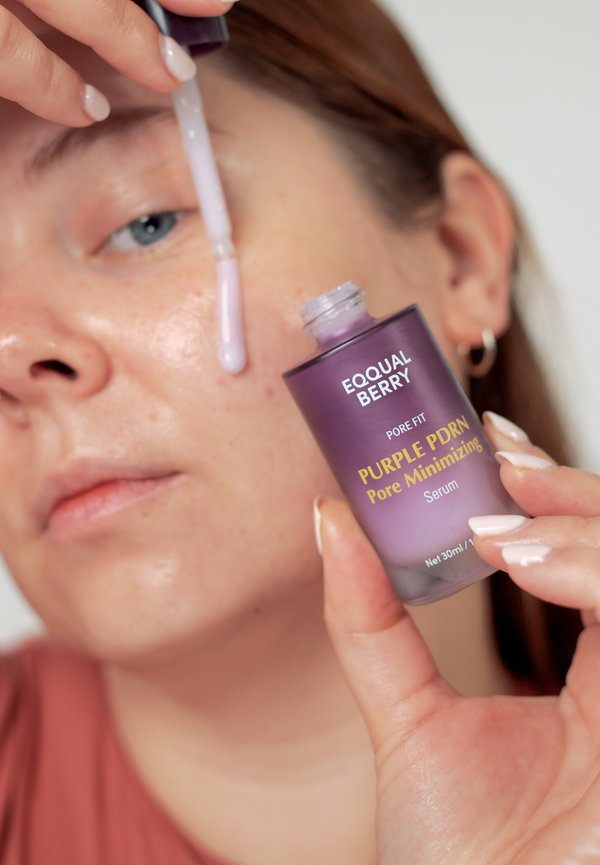 Osoba trzyma fioletową butelkę z etykietą "PURPLE PDRN Pore Minimizing Serum" i nakłada serum na policzek za pomocą pipety.