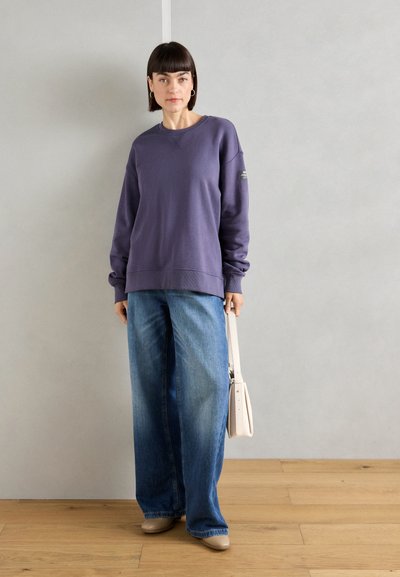 Sudadera morada de corte holgado con puños de canalé, combinada con vaqueros de pierna ancha de color azul. Detalle de pequeño logo en la manga. Zapatos beige y un bolso claro.