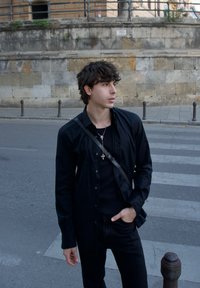 Fekete gombos ing fekete pulóver felett, fekete farmerral viselve. Ezüst keresztlánc és fekete váll táska. Kőfal a háttérben.