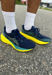 Zilās un dzeltenās sporta kurpes ir aprīkotas ar elpojošu augšējo daļu ar strukturētu dizainu, mīkstu zoli un kontrastējošiem šņorēm, kas sader ar baltām zeķēm.