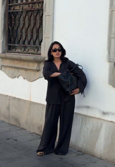Mujer vestida completamente de negro y con gafas de sol, sosteniendo una gran bolsa negra, de pie frente a una pared blanca con marcos de ventana de piedra.