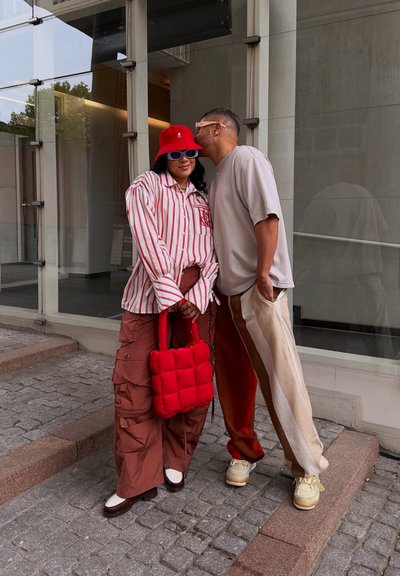 Dos personas posan al aire libre. Una de ellas lleva una camiseta de rayas rojas y blancas, pantalones cargo marrones y un sombrero de cubo rojo, sosteniendo una bolsa acolchada roja. La otra viste una camiseta beige y pantalones de rayas crema.