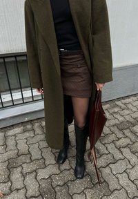 Personne portant un long manteau olive, un haut noir, une jupe marron à fines rayures, des bottes noires, portant un sac à franges en cuir marron foncé sur une allée extérieure pavée.