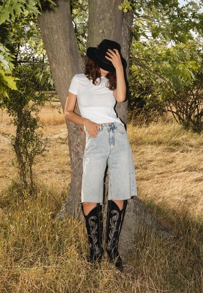 Camiseta blanca ajustada, culottes de mezclilla azul claro, botas vaqueras negras con bordados de plata, de pie contra un árbol en un campo de hierba.