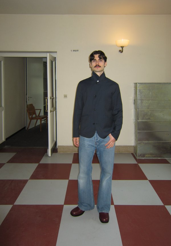 Άνδρας με σκούρα μαλλιά και μουστάκι στέκεται σε πλακιδωτό διάδρομο φορώντας σκούρο μπουφάν, ανοιχτό μπλε τζιν με καμπάνα και μπορντό παπούτσια.