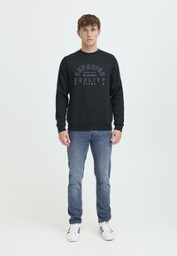 Black sweatshirt featuring "SUPERIOR QUALITY" printed in white, with a crew neck and ribbed cuffs. Styled with blue jeans and white trainers.
