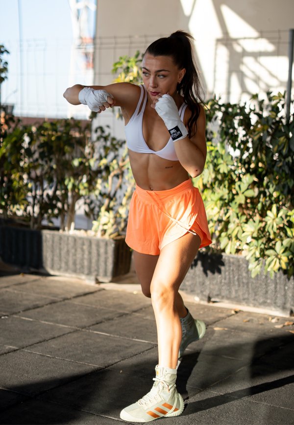 Femme en brassière blanche et short orange pratiquant des coups de boxe en plein air près de la verdure.