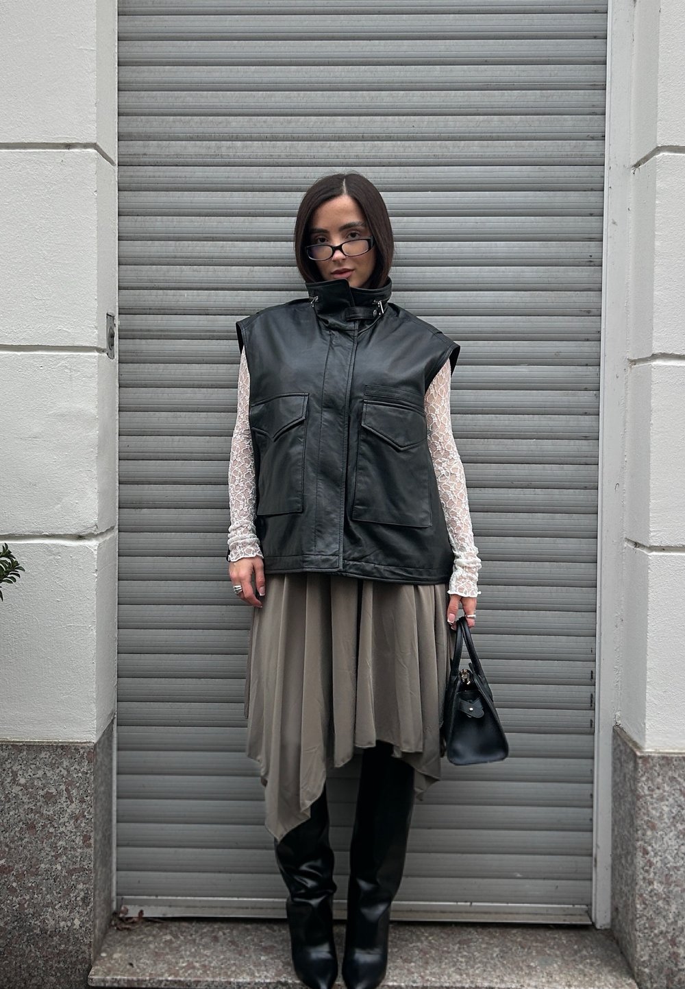 Czarna skórzana kamizelka bez rękawów z dwoma przednimi kieszeniami, nałożona na jasnobeżową asymetryczną spódnicę, w połączeniu z czarnymi kozakami do kolan i małą torebką.