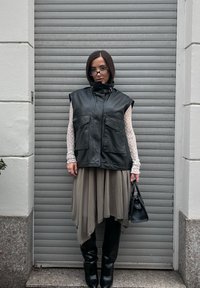 Czarna skórzana kamizelka bez rękawów z dwoma przednimi kieszeniami, nałożona na jasnobeżową asymetryczną spódnicę, w połączeniu z czarnymi kozakami do kolan i małą torebką.