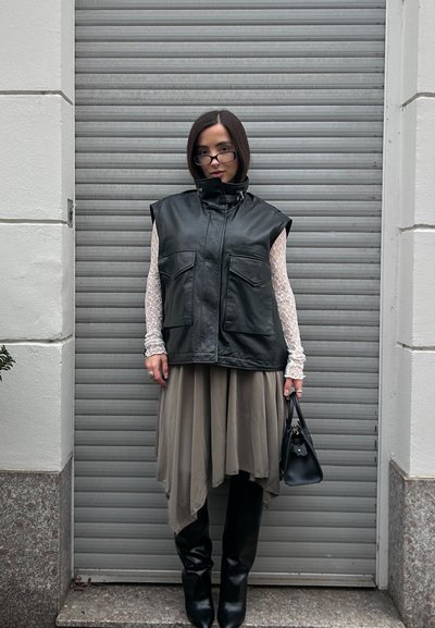 Chaleco negro sin mangas de cuero con dos bolsillos delanteros, superpuesto sobre una falda asimétrica beige claro, combinado con botas negras hasta la rodilla y un bolso pequeño.