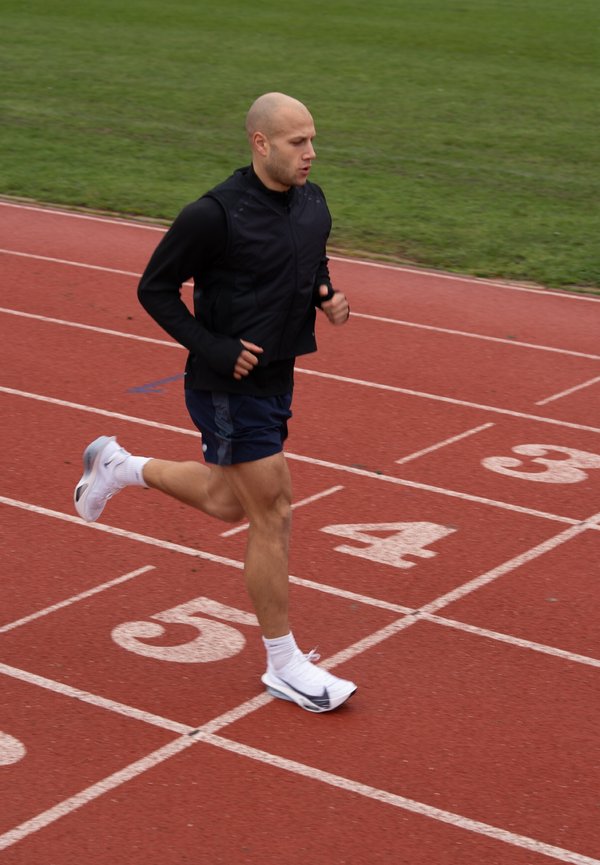Mann in schwarzer Weste und blauer Shorts läuft auf der roten Bahn in der Spur 5, im Hintergrund Gras.