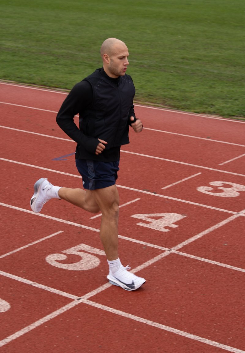 Man in zwart vest en blauwe korte broek rent op een rode baan in baan 5 met gras op de achtergrond.
