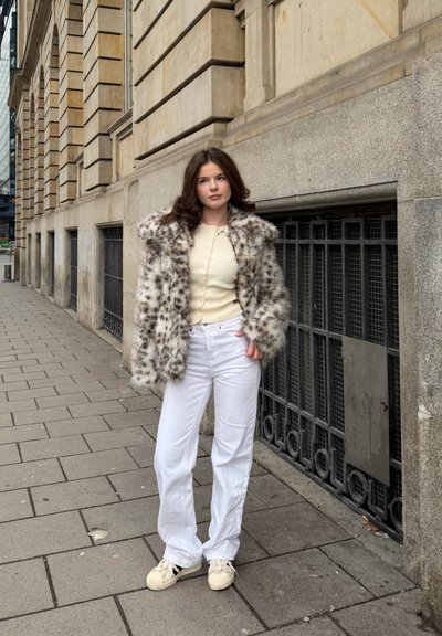 Joven mujer con un abrigo de piel sintética de manchas, top crema, pantalones blancos y zapatillas de deporte, de pie en la acera de una ciudad cerca de un edificio de piedra.