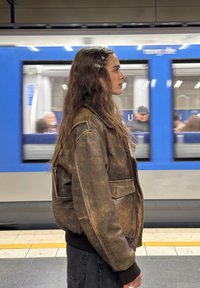 Smeđa kožna jakna kratkog kroja, nošena preko crne majice i tamnih hlača. Duga, valovita kosa s ukosnicama. Iza se kreće podzemni vlak.