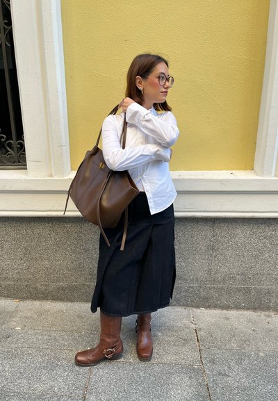 Bolso tote de cuero marrón con un diseño minimalista, combinado con una camisa blanca de botones y una falda azul marino, acompañado de botas de tobillo marrones.