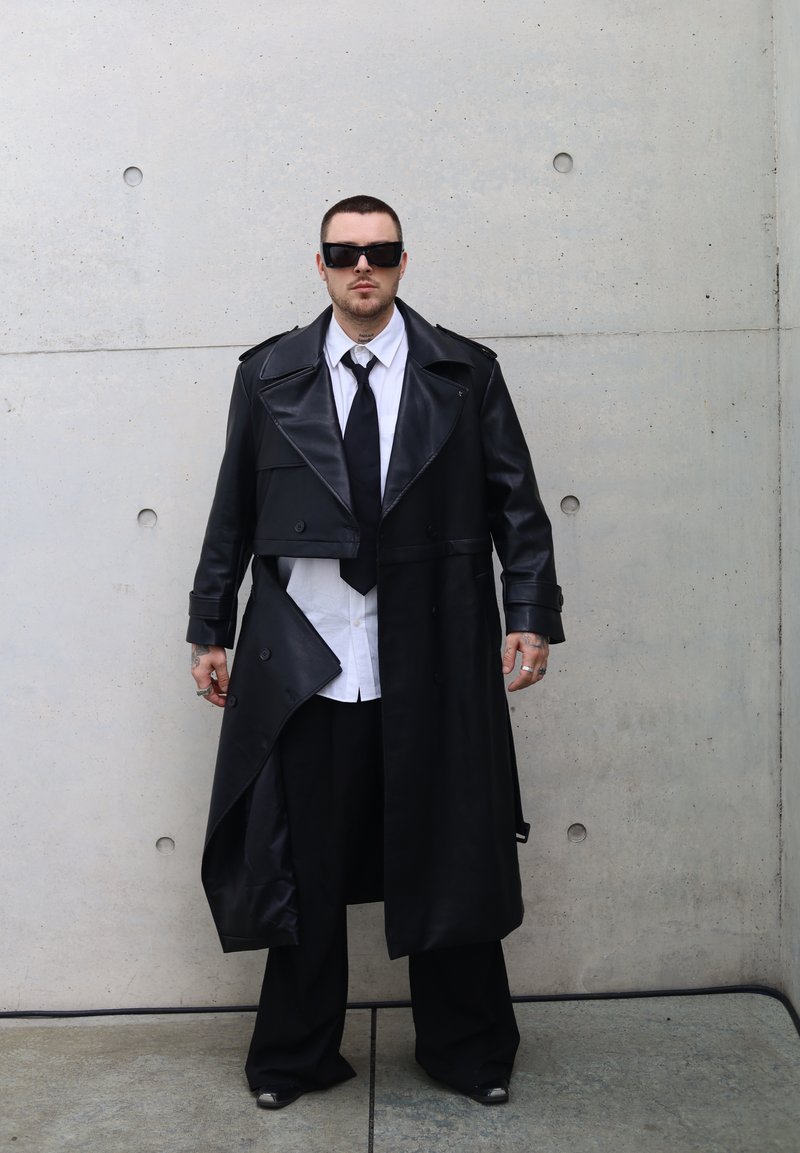 Trench en cuir noir avec des revers larges, design superposé, détails de boutons, porté sur une chemise blanche et une cravate noire, associé à un pantalon oversized.
