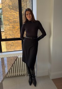 Col roulé noir, ceinturé à la taille, assorti à une jupe noire jusqu'aux genoux. Gants longs en cuir noir et bottes noires montantes à bout pointu.