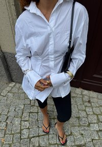 Valkoinen nappipaita, jossa on rypistynyt pinta, pitkät hihat, kultaiset yksityiskohdat, yhdistettynä mustiin leggings-housuihin ja flip flop -sandaleihin kivisellä pinnalla.