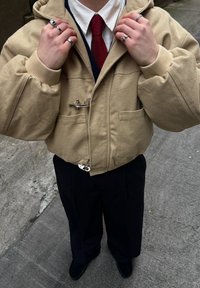 Bézs kapucnis dzseki cipzáras záródással és két elülső zsebbel, tengerészkék pulóver és fehér ing fölött viselve, sötét nadrággal párosítva.