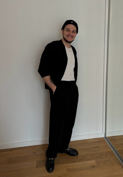 Cárdigan negro sobre una camiseta blanca, pantalones negros y zapatos negros con hebilla. El fondo es una pared blanca lisa.