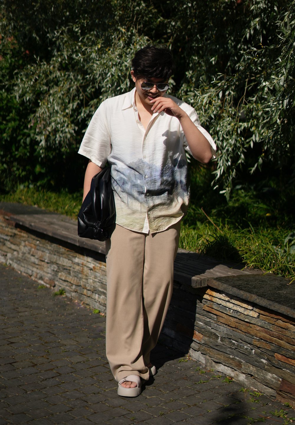 Ανοιχτόχρωμο πουκάμισο κοντομάνικο με σχέδιο τοπίου, μπεζ φαρδιά παντελόνια και υπερμεγέθη μαύρη τσάντα; άτομο που φορά σανδάλια και γυαλιά ηλίου.