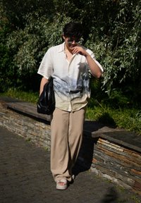 Ανοιχτόχρωμο πουκάμισο κοντομάνικο με σχέδιο τοπίου, μπεζ φαρδιά παντελόνια και υπερμεγέθη μαύρη τσάντα; άτομο που φορά σανδάλια και γυαλιά ηλίου.