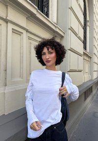 Vrouw met krullend haar, gekleed in een lange witte shirt en een donkere broek, die staat en een zwarte tasband vasthoudt op een stoep in de stad.
