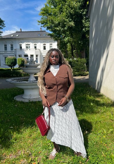 Chaleco marrón de rayas finas sobre un vestido de encaje blanco, sosteniendo un bolso burdeos. El fondo presenta una fuente y vegetación.