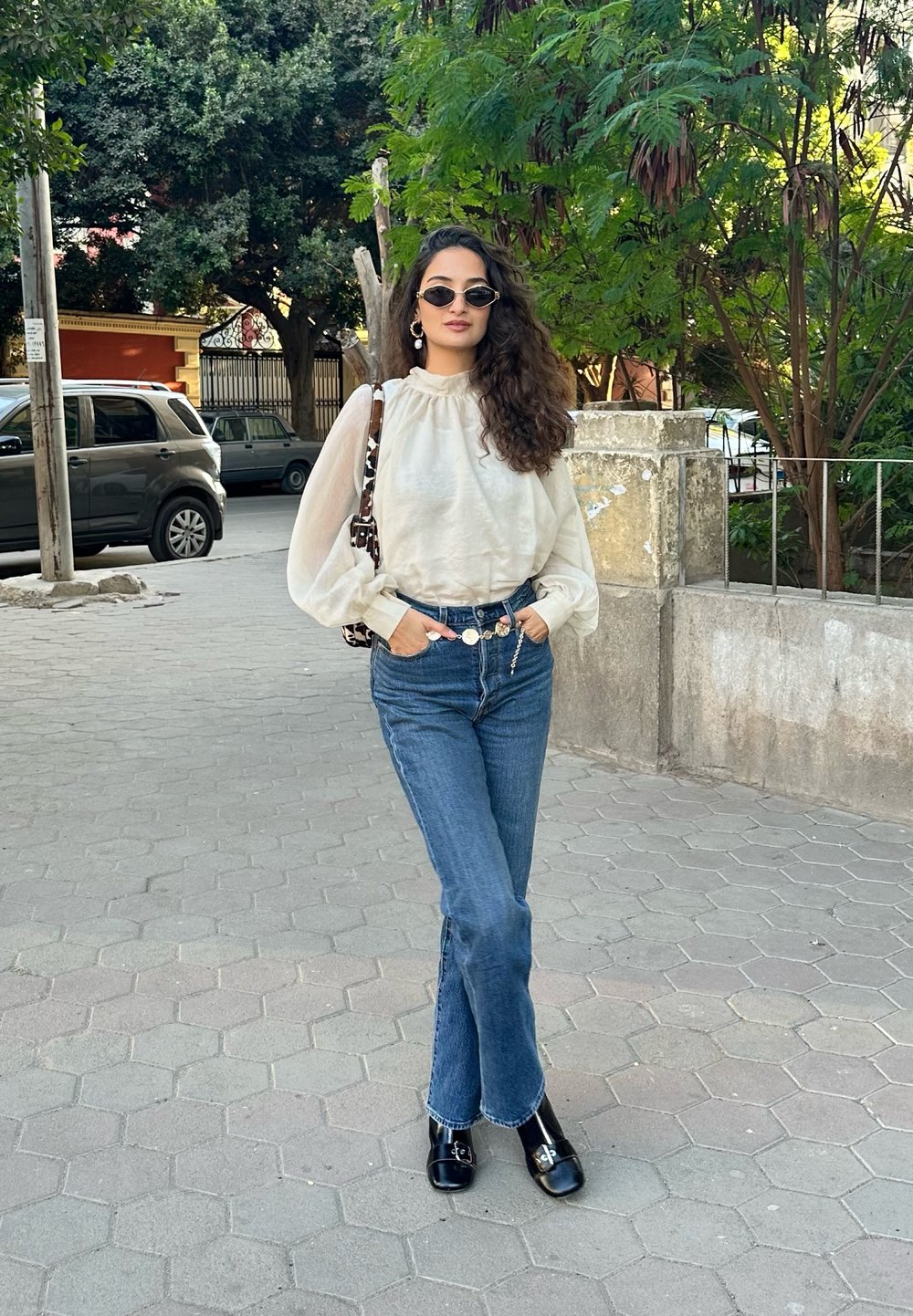 Blouse beige clair à manches longues avec un col froncé ; jean bleu taille haute évasé ; bottines noires ; sac à imprimé animalier. Décor urbain.