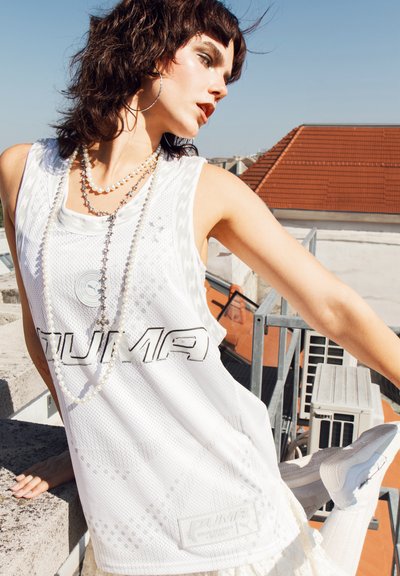 Top sin mangas de malla blanca con gran logo de "PUMA", escote redondo y patrón geométrico; combinado con collares de perlas en capas. El fondo presenta techos.